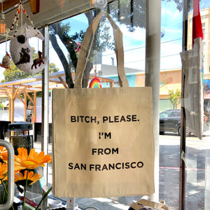 Simple beige canvas bag with two straps hanging in a window with "Bitch, please. I'm from San Francisco" in bold, black, all cap text.
